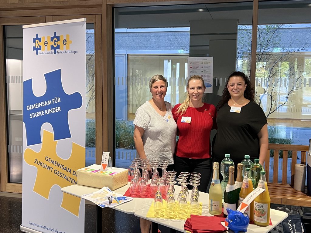 Ein Tisch mit Gläsern, Flaschen und eine Spendenbox, hinter dem drei Damen vom Förderverein ReGe stehen. Daneben ein Rollup-Poster mit dem Logo des Fördervereins.
